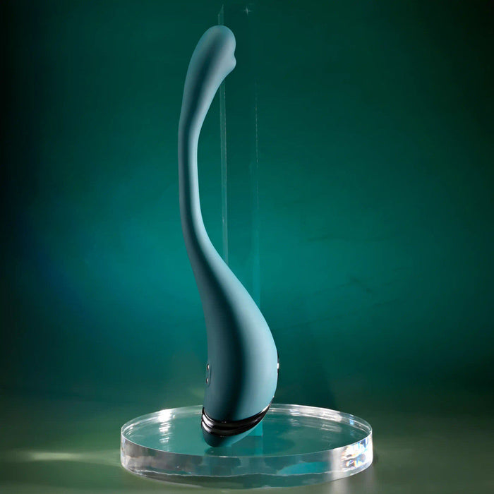 The Pinpoint Perfection Flexible Slim Vibrator, in teal with a curved modern design, stands upright on a clear round stand against a green gradient background.