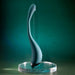 The Pinpoint Perfection Flexible Slim Vibrator, in teal with a curved modern design, stands upright on a clear round stand against a green gradient background.