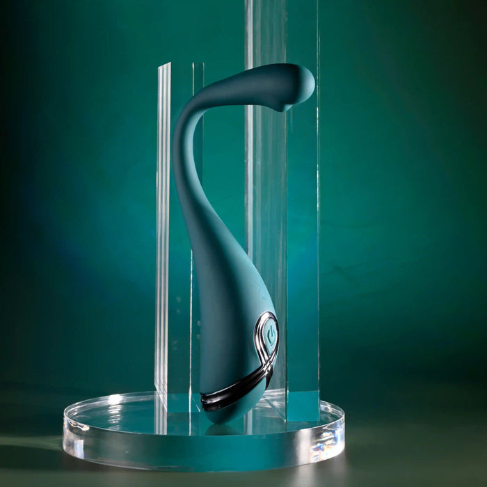 The Pinpoint Perfection Flexible Slim Vibrator, a sleek teal device with a curved design, stands upright on a clear round platform amid vertical acrylic panels, set against a green gradient background.