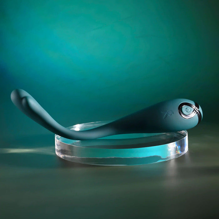 The Pinpoint Perfection Flexible Slim Vibrator, featuring a sleek curved teal design and circular control end, rests on a transparent round stand against a teal gradient background.