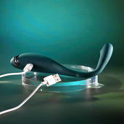 The Pinpoint Perfection Flexible Slim Vibrator, in dark green with a USB charging cable attached, rests on a clear circular stand against a teal and green background.