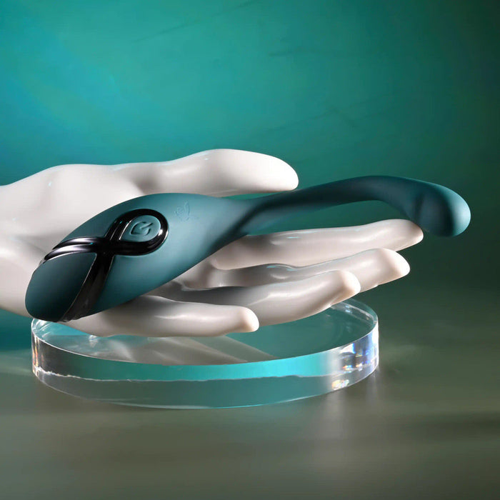 A sculpted white hand holds the Pinpoint Perfection Flexible Slim Vibrator, a sleek teal device with a button and curved design, resting on a clear round stand against a matching teal background.
