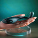 A realistic mannequin hand with manicured nails holds the Pinpoint Perfection Flexible Slim Vibrator—a sleek, dark teal, curved, bendable device—above a clear round platform against a soft teal background.