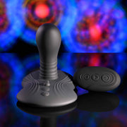 The Playboy Sittin' & Spinnin' Hands Free G-Spot Vibrating Grinder Pad, a gray silicone toy with a rounded top and textured base, sits next to its black remote on an abstract blue, red, and purple background.
