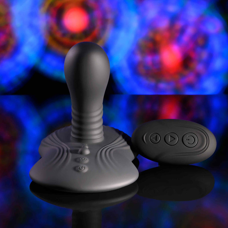 The Playboy Sittin' & Spinnin' Hands Free G-Spot Vibrating Grinder Pad, a gray silicone toy with a rounded top and textured base, sits next to its black remote on an abstract blue, red, and purple background.