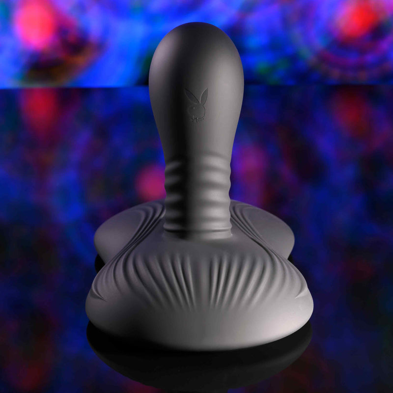 The Playboy Sittin' & Spinnin' Hands Free G-Spot Vibrating Grinder Pad, a gray silicone adult toy with ribbed texture and rabbit logo, is displayed upright on a reflective surface against a vibrant blue, red, and purple abstract background.