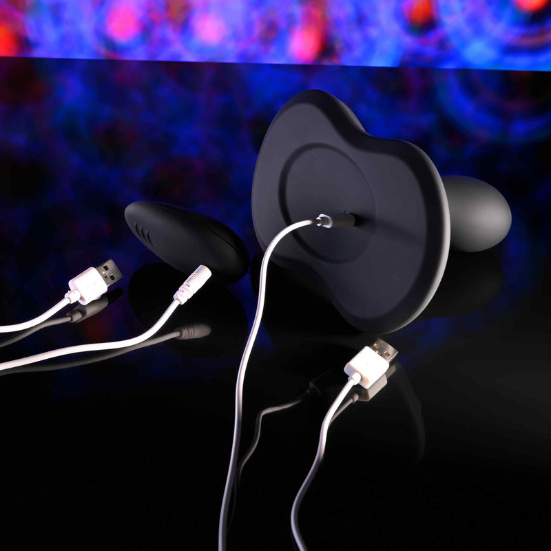 The Playboy Sittin' & Spinnin' Hands Free G-Spot Vibrating Grinder Pad and a black silicone rotating penetrator, both with white USB charging cables, rest on a glossy black surface with a colorful abstract pattern in the background.