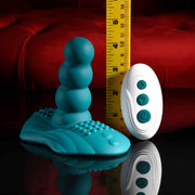 The Playboy Have a Seat Remote Control Grinding Vibrator Pad, made of teal body-safe silicone with a beaded shaft and textured suction cup base, is shown next to a white remote and yellow measuring tape against a red tufted background.