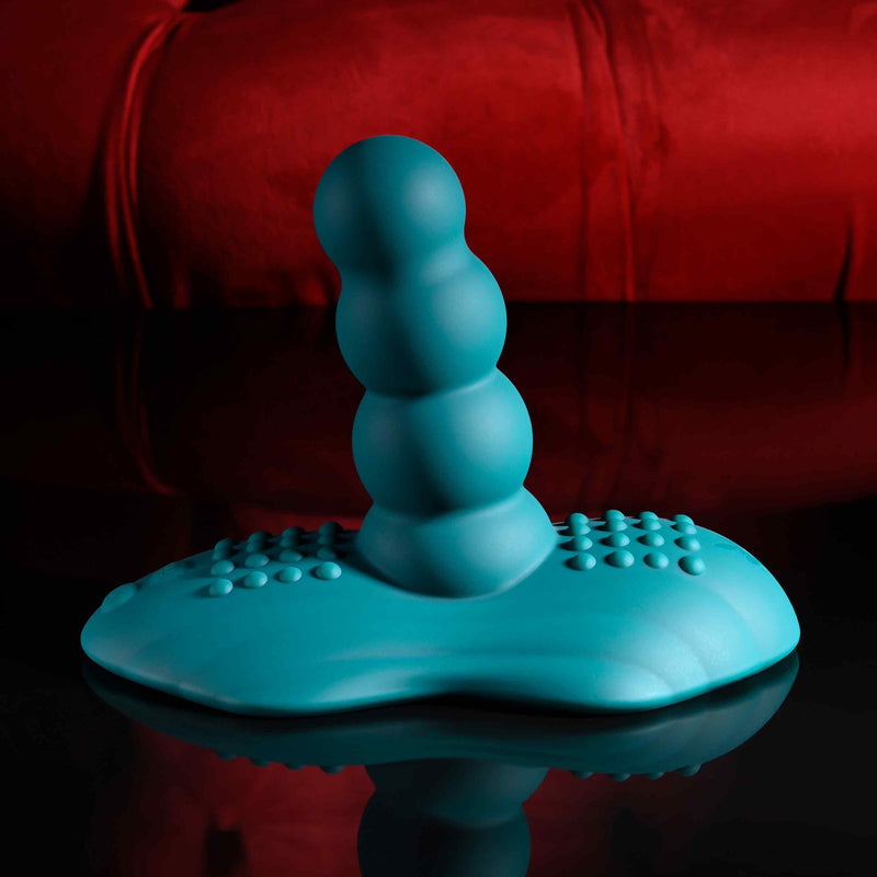 The Playboy Have a Seat Remote Control Grinding Vibrator Pad, made from body-safe silicone with a textured blue design and ridged suction base, rests on a reflective surface against a red, blurred backdrop.