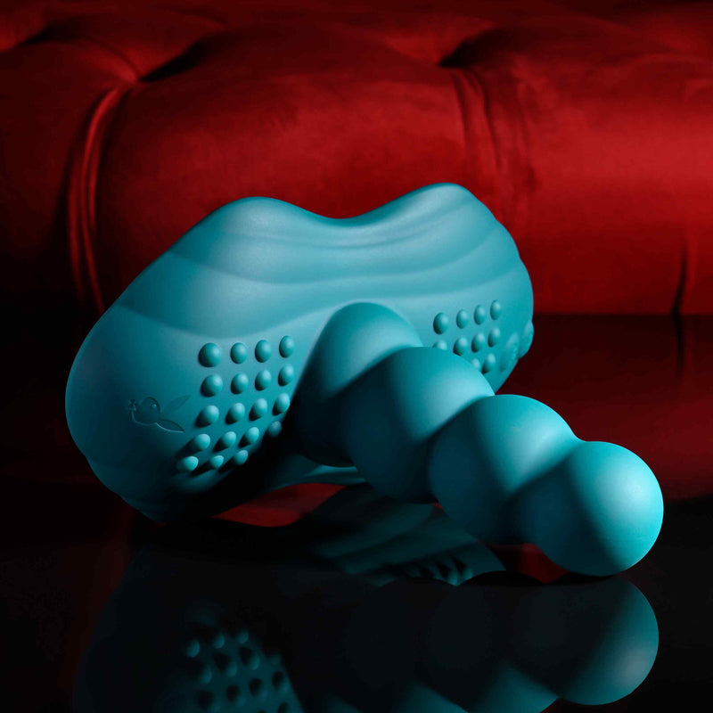 The Playboy Have a Seat Remote Control Grinding Vibrator Pad, made from body-safe silicone with a curved, ridged design and suction cup base, sits on a reflective black surface in front of a plush red cushion.