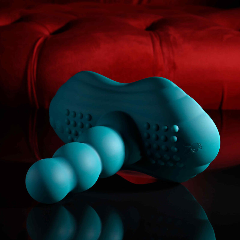 The Playboy Have a Seat Remote Control Grinding Vibrator Pad, made of teal, textured, body-safe silicone with a beaded shaft, nubs, and suction cup base, is displayed on a reflective black surface against a plush red background.