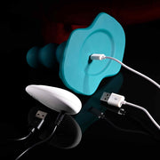 The Playboy Have a Seat Remote Control Grinding Vibrator Pads, in teal and white body-safe silicone, are being charged with USB cables on a dark reflective surface.