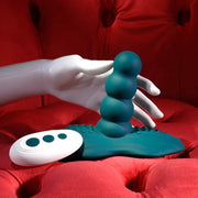 A white mannequin hand holds the Playboy Have a Seat Remote Control Grinding Vibrator Pad in teal, made from body-safe silicone, with its sleek remote resting on a red tufted velvet surface.