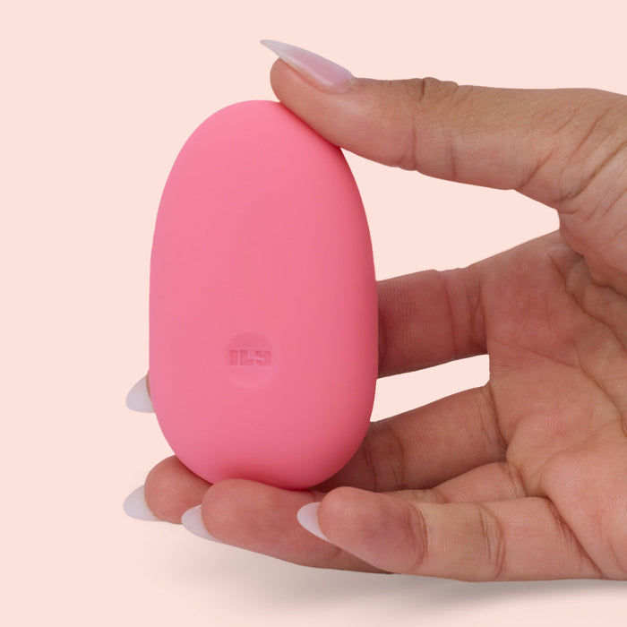 A hand with manicured nails holds the Ily Squishy Silicone Pebble Vibrator, a small, smooth, pink device made of body-safe silicone, against a light pink background.
