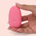 A hand with manicured nails holds the Ily Squishy Silicone Pebble Vibrator, a small, smooth, pink device made of body-safe silicone, against a light pink background.
