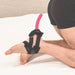 A person in black shorts is on their hands and knees on a bed, showcasing the Pivot Flex Foot and Ankle Strap On Harness by Sportsheets, with a pink dildo extending from it. The background reveals a beige headboard and white sheets, creating an ideal setting for solo or couple play.