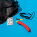 A black bag, cat-eye sunglasses, a sheet of stickers, lip gloss, and the Romp Hype Rechargeable Silicone G-Spot Vibrator in red and yellow are arranged on a bright blue background.