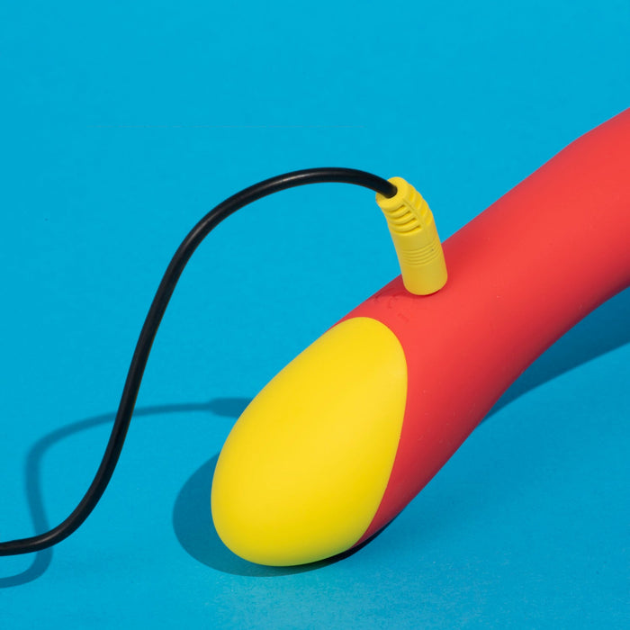 The Romp Hype Rechargeable Silicone G-Spot Vibrator, shown in red and yellow with a rounded end, is being charged with a black cable against a bright blue background.