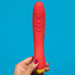 A hand with painted nails holds the Romp Hype Rechargeable Silicone G-Spot Vibrator upright against a solid blue background, highlighting its red and yellow design for intimate G-spot exploration.