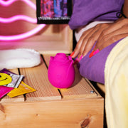 The Romp Rose Pleasure Air Clitoris Stimulator - Pink sits on a wooden bedside table beside condom packets, with a hand on a purple bedsheet. Neon pink lights and cozy bedding complete the relaxed vibe.