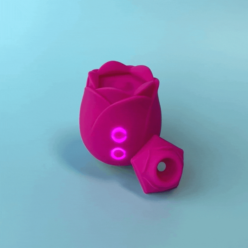 The Romp Rose Pleasure Air Clitoris Stimulator - Pink, a rechargeable silicone toy with glowing indicator lights, sits on a light blue surface next to its matching cap with a central hole, showcasing innovative pleasure air technology.
