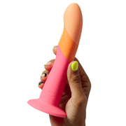 A hand with colorful nail polish holds the Romp Dizi Ultra Smooth 7 Inch Dildo With Suction Cup, a brightly colored, two-tone silicone toy with a flared base designed for sexual pleasure.