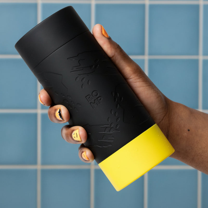 A hand with painted nails holds the black Romp Dash Discreet Stroker for Beginners, its yellow, textured design evoking a stylish travel mug embossed with "ROMP," set against a blue tiled wall.
