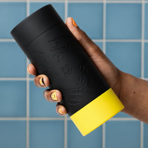 A hand with painted nails holds the black Romp Dash Discreet Stroker for Beginners, its yellow, textured design evoking a stylish travel mug embossed with "ROMP," set against a blue tiled wall.