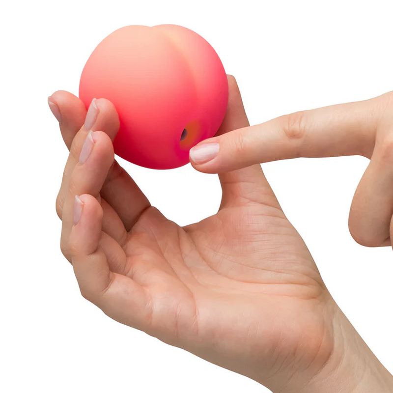 Hand holding a compact, soft silicone peach-shaped clitoris stimulator with a finger pointing to its opening.