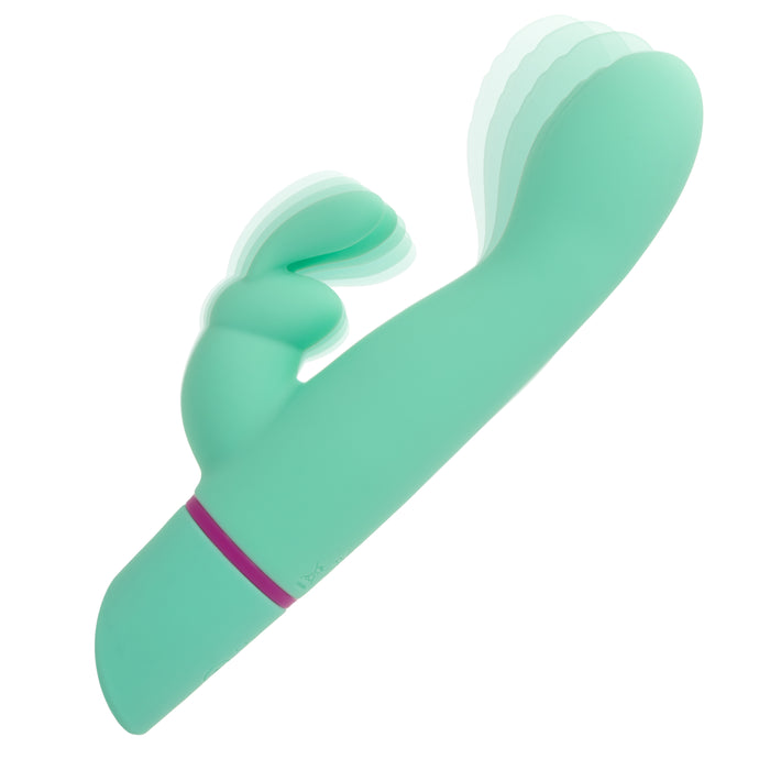 The Love Bunny Rockin’ G Triple Motor Rabbit Vibrator, shown in turquoise with a pink-accented handle, features a curved shaft and smaller extension for dual stimulation. Blurred motion lines indicate its triple motors vibrating against a white background.