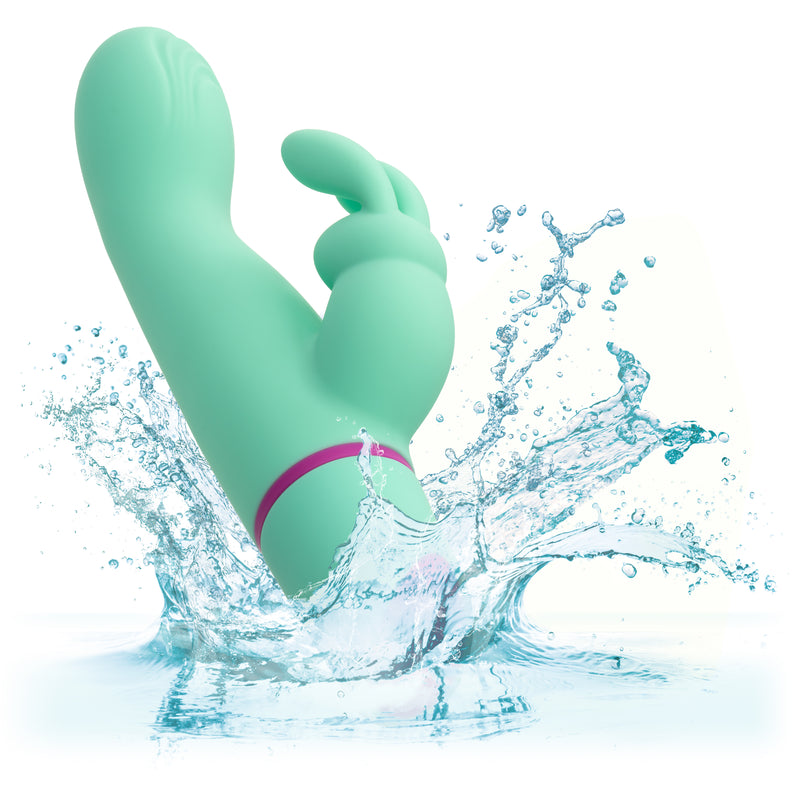 The mint green Love Bunny Rockin’ G Triple Motor Rabbit Vibrator, featuring a curved tip and smaller extension, splashes into clear water, showcasing its waterproof design and suitability for wet environments.