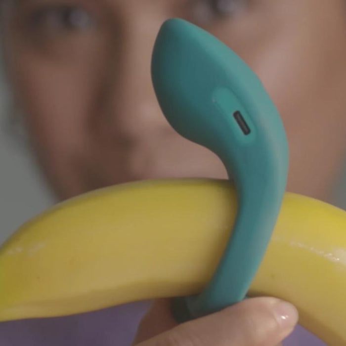 A person holds a yellow banana next to the Dame Hug Adjustable Vibrating Cock Ring for Couples, comparing their shapes. The teal cock ring, featuring a small charging port, is held near the face, which appears out of focus.