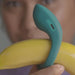 A person holds a yellow banana next to the Dame Hug Adjustable Vibrating Cock Ring for Couples, comparing their shapes. The teal cock ring, featuring a small charging port, is held near the face, which appears out of focus.