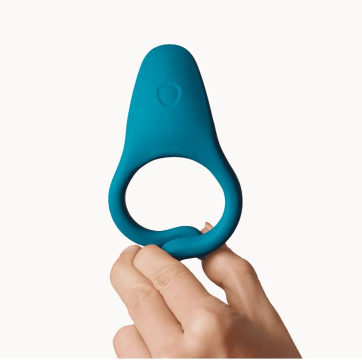A hand holds up the Dame Hug Adjustable Vibrating Cock Ring for Couples, featuring a blue silicone design with a wider, flat top, against a plain white background.