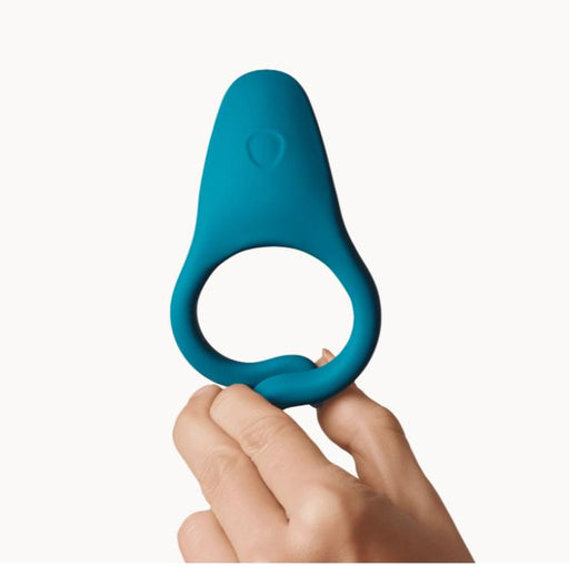 A hand holds up the Dame Hug Adjustable Vibrating Cock Ring for Couples, featuring a blue silicone design with a wider, flat top, against a plain white background.