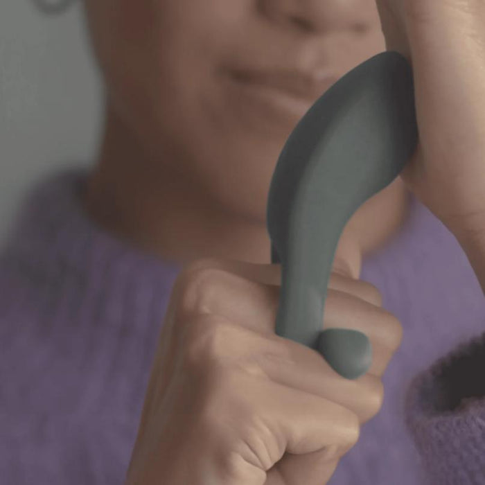 A person wearing a purple sweater holds the Dame Hug Adjustable Vibrating Cock Ring for Couples in their hand, thumb on its small button. Their partially visible face is blurred in the background.