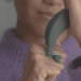 A person wearing a purple sweater holds the Dame Hug Adjustable Vibrating Cock Ring for Couples in their hand, thumb on its small button. Their partially visible face is blurred in the background.