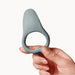 A hand holds the Dame Hug Adjustable Vibrating Cock Ring for Couples, a soft grey silicone ring with a raised button, against a plain white background.