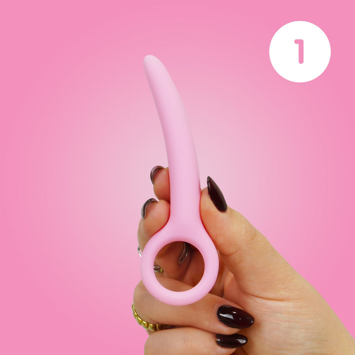 A hand with dark nail polish holds a Skins Care Curved Vaginal Dilator (Small), made of pink medical-grade silicone with a rounded ring handle, against a pink backdrop with a white "1," supporting pelvic health. Part of 3-piece set (Small/Medium).