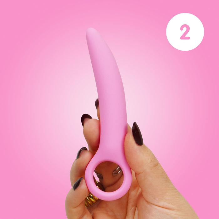 A hand with dark nail polish holds a small pink Skins Care Curved Vaginal Dilator from the 3-Piece Set against a pink gradient background. A white circle with the number 2 appears in the upper right corner.