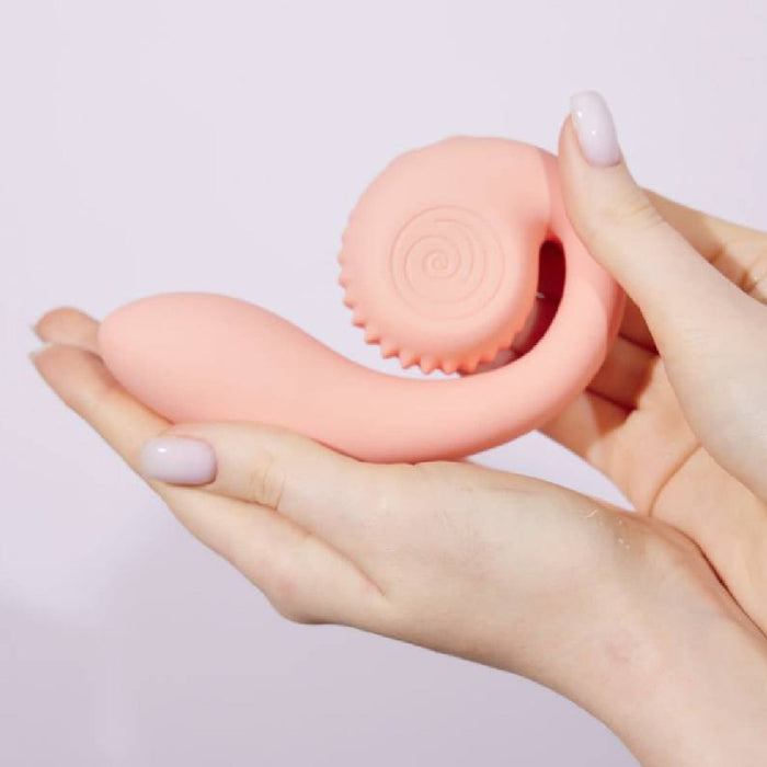 A person with neatly manicured nails holds the Snail Gizi Compact Ultra Powerful 2 Motor Dual Stimulating Vibrator in pink, designed by Snail Vibe for G-spot stimulation. The vibrator features a bulbous tip and a textured, spiral design on one end. The background is neutral and light-colored.