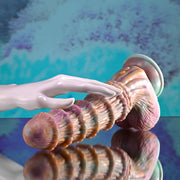 The Fantasy Ultra Textured Vibrating Remote Control Creature Dildo, featuring a sculpted pastel design and ribbed texture, rests on a reflective surface as a mannequin-like hand gently touches it. The blurred background glows blue and purple.