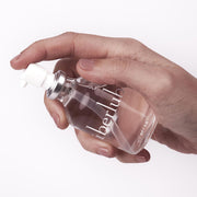 A hand presses the pump of a clear UberLube Premium Silicone Lubricant 50 ml bottle, featuring a silver cap and white nozzle, against a white background.