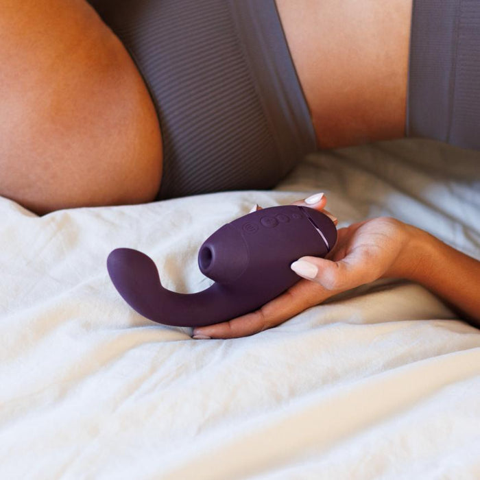 A person lying on a bed, dressed in a grey ribbed garment, holds the Womanizer Next Duo Pleasure Air + Vibration Blended Orgasm Stimulator against light bedding.