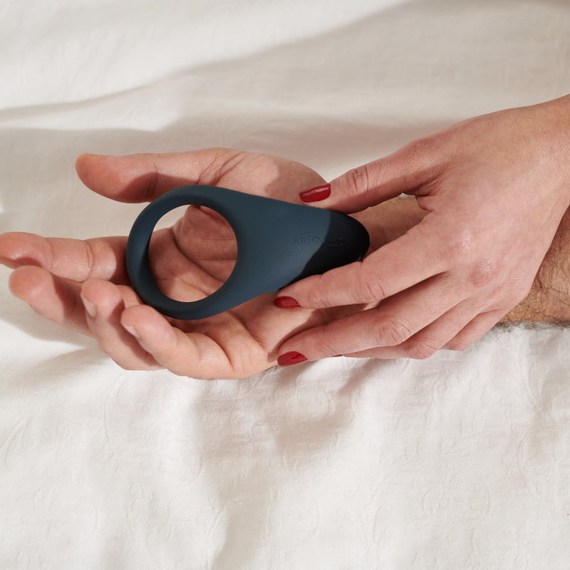 Dark blue silicone We-Vibe Verge vibrating cock ring held in two hands on a white sheet.