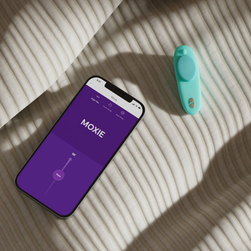 A teal We-Vibe Moxie + Hands-Free Remote or App Controlled Panty Vibrator rests on striped beige fabric beside a smartphone showing the purple MOXIE app screen, bathed in sunlight that highlights this discreet wearable vibrator.