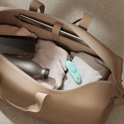 A beige bag is open on a light carpet, revealing folded clothes, a silver water bottle, a smartphone, a teal We-Vibe Moxie+ Hands-Free App Controlled Panty Vibrator, and a laptop inside.
