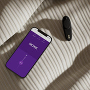 A smartphone displaying a purple "MOXIE" screen sits beside the We-Vibe Moxie + Hands-Free Remote or App Controlled Panty Vibrator in black, both resting on a light-colored, striped fabric surface.