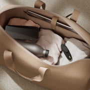 A tan leather duffel bag with an open top, showing neatly packed clothes, a silver water bottle, a black smartphone, a slim laptop, and a discreetly tucked We-Vibe Moxie+ hands-free app controlled panty vibrator in black.