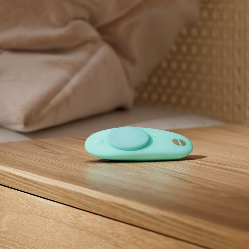 A teal We-Vibe Moxie+ hands-free remote or app-controlled panty vibrator rests on a wooden nightstand beside a bed with beige bedding.